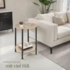 A round side table with two shelves is placed next to a sofa in a living room. Náhled obrázku produktu