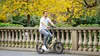A man rides an electric bicycle along a path next to a stone balustrade with yellow trees in the background. Produktbild-Vorschau