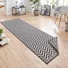 A long runner rug with a black and white chevron pattern is laid out on a light wood floor in a room with a window and furniture. Voir les images du produit