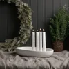 A white ceramic candle holder with four white candles is placed on a gray linen tablecloth. A festive wreath and a potted evergreen plant are visible in the background. Produktbild-Vorschau