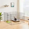 A dog is inside a black wire playpen with a dog bed. The playpen is set up on a wooden floor in a room with a window and a shelf on the wall. Produktbild-Vorschau