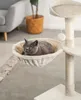A cat is resting in a beige hammock attached to a cat tree, with a chair and a potted plant in the background. Produktbild-Vorschau