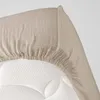 A close-up view of a beige muslin fitted sheet draped over a white mattress. Náhled obrázku produktu