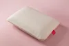 A white pillow with a textured pattern and gold trim is placed on a light pink background. A small red tag with a white cloud logo is attached to the corner of the pillow. Produktbild-Vorschau