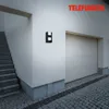 A black wall-mounted light fixture is shown next to a garage door and outdoor stairs. Produktbild-Vorschau