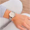 A person wearing white pants and a striped shirt is shown wearing a silver watch on their wrist. Produktbild-Vorschau