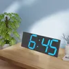 A digital clock with blue digits is placed on a wooden table next to a potted plant. Náhled obrázku produktu