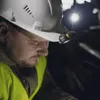 A close-up of a construction worker wearing a hard hat with a headlamp attached, working in a dark environment. Produktbild-Vorschau