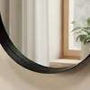 A close-up of a round black-framed mirror reflecting a window with white curtains and a potted plant. Produktbild-Vorschau