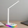A white LED desk lamp with a colorful RGB base is placed on a wooden desk next to an open book and a laptop. Produktbild-Vorschau