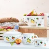 A collection of white enamelware kitchen items decorated with fruit patterns, displayed on a wooden shelf with a loaf of bread and fresh fruit in the background. Produktbild-Vorschau