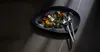 A salad is served on a dark, irregular plate with two serving spoons on the side, set against a dark, textured surface with dramatic lighting. Anteprima dell’immagine prodotto
