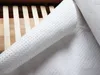 A close-up view of a white mattress protector with raised dots, partially unrolled on a wooden bed frame. Produktbild-Vorschau