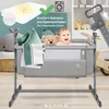 A baby is in a grey bassinet next to a teddy bear and pillows. The bassinet is on a wooden floor with a basket and a toy in the background. There is text and graphics in the upper left corner. Produktbild-Vorschau