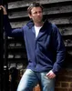 A man leaning against a wooden wall, wearing a navy blue fleece jacket and jeans. Produktbild-Vorschau
