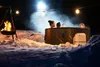 People relaxing in a hot tub in the snow at night, with a campfire and string lights. Produktbild-Vorschau