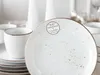 A close-up view of white speckled ceramic plates and cups, with a label on the plate. Náhľad obrázku produktu
