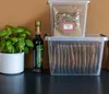A clear plastic storage box with a lid, containing several brown paper bags, is stacked on top of another identical box. A bottle of dark liquid, a potted green plant, and a wooden object are placed to the left of the boxes. Anteprima dell’immagine prodotto