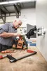 An older man is working on a wooden birdhouse on a workbench, with tools and a tablet mounted on a wall nearby. Podgląd zdjęcia produktu