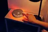 A hand is pressing a button on a baby sleep aid device on a nightstand next to a lamp. Náhľad obrázku produktu