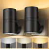 Two black LED plug-in night lights are shown mounted on a wall, with a warm yellow light cast upwards. Below the main image, three smaller panels display the lights emitting different shades of light: white, warm white, and yellow. Produktbild-Vorschau