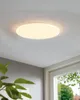 A white, circular ceiling lamp is mounted on a white ceiling. The lamp is illuminated, casting a soft glow. In the foreground, out of focus, is a green plant and a window with trees visible outside. Produktbild-Vorschau