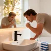 A man brushing his teeth in a modern bathroom with a sink and bathtub visible. Produktbild-Vorschau