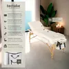 A white massage table is set up in a room with a plant and candles in the background. Náhled obrázku produktu