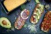 A top-down view of three gourmet hot dogs with various toppings, served on a dark slate surface, with a block of cheese and avocado halves nearby. Náhled obrázku produktu