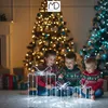 Three children are sitting on the floor in front of Christmas trees, looking at illuminated gift decorations. Podgląd zdjęcia produktu