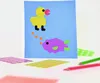 A craft project featuring a blue background with a yellow duck and a purple fish made of paper shapes. Several sheets of colorful dot stickers and two colored pencils are scattered around the base of the craft. Produktbild-Vorschau