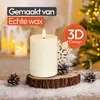 A cream-colored LED candle with a flame sits on a wooden slice, surrounded by snow and pinecones, with a Christmas tree in the background. Produktbild-Vorschau