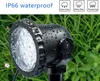 A black LED garden spotlight covered in water droplets, set against a background of green grass and some small pink flowers. Produktbild-Vorschau
