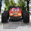 A remote-controlled off-road car with large tires is shown from the front, with a blurred background of trees and a road. Anteprima dell’immagine prodotto