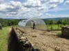 A greenhouse is set up in a rural, hilly landscape with trees and vegetation under a cloudy sky. Produktbild-Vorschau