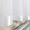 A close-up view of white sheer curtains with a satin hem, draped over a windowsill with a radiator below. Produktbild-Vorschau