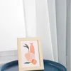 A framed picture with a modern abstract design of vases and fruit sits on a dark blue tray, with sheer grey and white curtains in the background. Produktbild-Vorschau