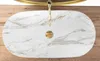 A top-down view of a white oval countertop sink with gold veining and a gold faucet, placed on a wooden surface. Náhľad obrázku produktu