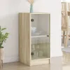 A wooden cabinet with glass doors is shown in a room with a plant and a chair in the background. Náhľad obrázku produktu