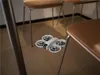 A small white drone is hovering near the legs of chairs in a room. Anteprima dell’immagine prodotto