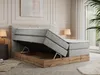 A bedroom scene featuring a king-size bed with its storage compartment open, revealing the wooden frame and storage space. A bedside table with a lamp is visible on the left, and a window with curtains and a plant is on the right. Produktbild-Vorschau