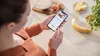 A person holding a smartphone with a recipe on it, in a kitchen setting with ingredients visible. Produktbild-Vorschau