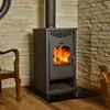 A wood-burning stove with a fire burning inside, placed in a room with a stone wall and wooden floor. A chair and a basket of firewood are also visible. Anteprima dell’immagine prodotto