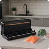 A vacuum sealer machine is shown on a kitchen counter with sealed bags of food. Podgląd zdjęcia produktu