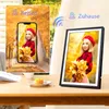 A digital photo frame is shown on a table, with a smartphone displaying the same image in the background, illustrating photo transfer. Produktbild-Vorschau