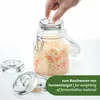 A hand is placing a glass lid on a jar filled with fermented food, with other glass weights in the foreground. Produktbild-Vorschau