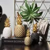 A collection of decorative pineapple ornaments displayed on a shelf with a book and a framed picture. Produktbild-Vorschau