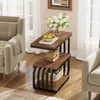 A narrow side table with a rustic wood finish and black metal frame, placed between two armchairs in a living room setting. It is decorated with a vase of flowers, a bowl of fruit, and a small decorative radio. Produktbild-Vorschau