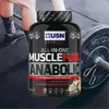 A person's legs and a weightlifting barbell are visible in the background, with a large tub of USN Muscle Fuel Anabolic protein powder in the foreground. Produktbild-Vorschau