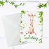 A greeting card with a giraffe illustration and jungle leaves, placed next to a white envelope on a white wooden surface. Produktbild-Vorschau